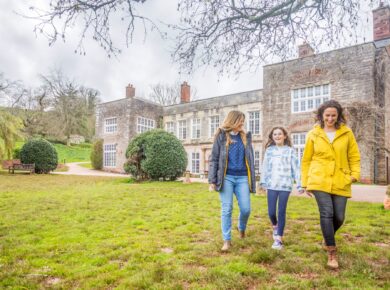 Two Women And A Child Walking In Front Of Cockington Court'S Manor House.