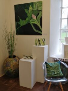 A Selection Of Art From The Flora Whispers Exhibition. There Is A Large Painting On The Wall, Below It Is Two Pedestals With Glass Work On Them. To The Right Is A Small Chair With A Green Cushion, And To The Left A Large Plant Pot With Grass Like Flora In In.  
