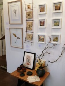 A Selection Of Art From The Flora Whispers Exhibition. There Are Art In Frames Hung On The All And A Small Wooden Table Below With A Selection Of Art And Vases. 